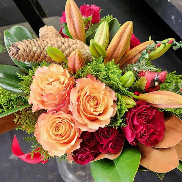 Peach and red roses with orange lilies, pinecones, and a red cardinal pick in a clear vase.