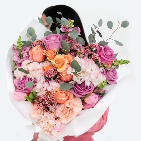 Large bouquet of pink, peach, and lavender flowers wrapped in white paper, held by a person in pink.