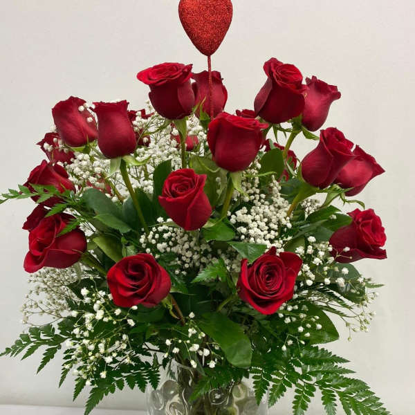 Red roses arranged in a clear glass vase with baby's breath and a heart pick