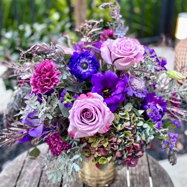 Purple and pink floral arrangement with roses, carnations, and hydrangeas in a gold vase