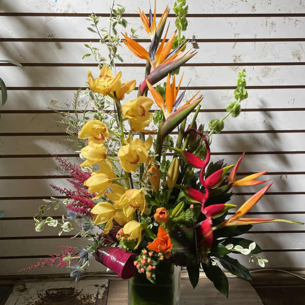 Tall tropical arrangement with yellow orchids and orange blooms in a green leaf-lined glass vase