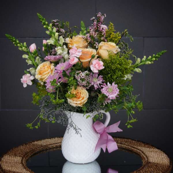 Peach roses and pink flowers in a white pitcher vase with a pink ribbon