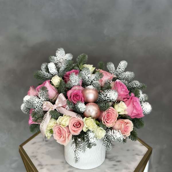 Pink and cream roses arranged in a white vase with frosted evergreen accents.