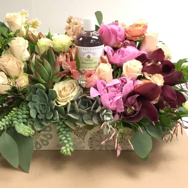 Mixed floral arrangement with roses, orchids, succulents, and a bath oil bottle in a decorative box