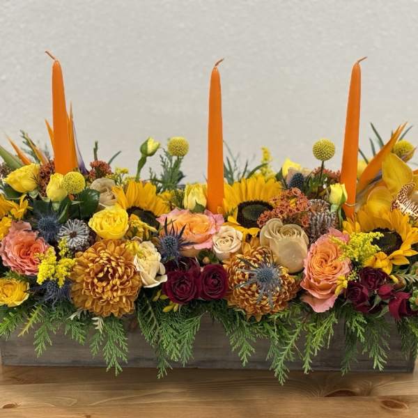 Long floral centerpiece with orange candles and mixed yellow, pink, and burgundy blooms