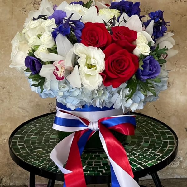 Red, white, and blue floral arrangement in a round box with ribbon