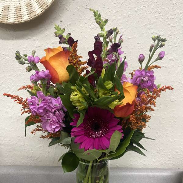 Mixed bouquet with orange roses, purple blooms, and a magenta gerbera in a glass vase