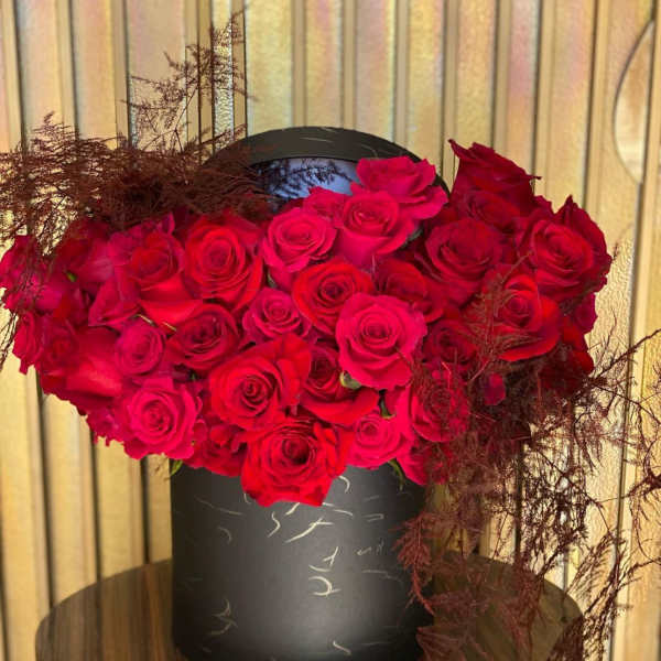 Red roses arranged in a black round box with dark feathery accents