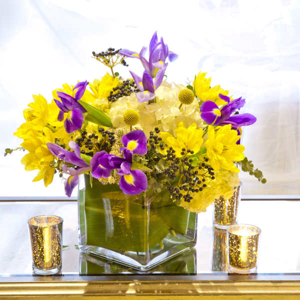 Purple irises and yellow flowers in a square glass vase with candles