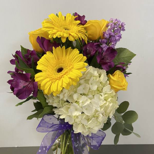 Bouquet of yellow gerbera daisies, roses, purple flowers, and white hydrangea in a glass vase