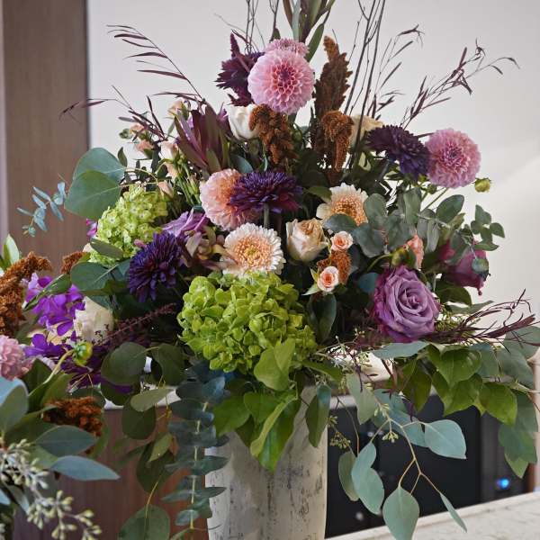Large mixed arrangement of purple, pink, and green flowers with tall branches in a stone urn vase
