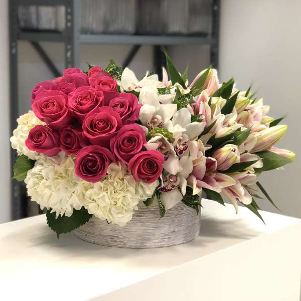 Low arrangement of hot pink roses, white hydrangeas, orchids, and pink lilies in a round white container.
