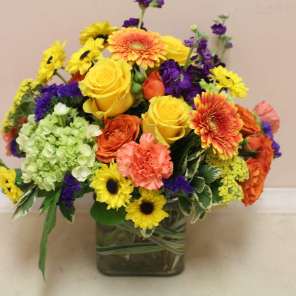 Mixed bouquet of yellow, orange, purple, and green flowers in a square glass vase