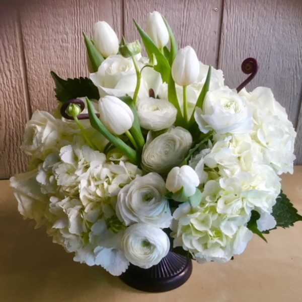 White floral arrangement with tulips, roses, ranunculus, and hydrangeas in a dark vase