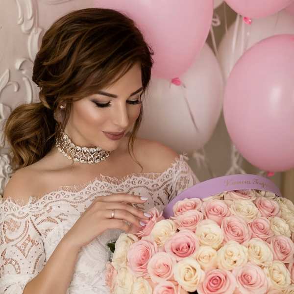 Woman in a white lace dress holding a box of pink and cream roses with balloons behind her
