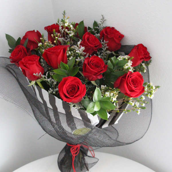 Hand-tied bouquet of red roses with white filler flowers wrapped in black mesh, with rose petals on a table