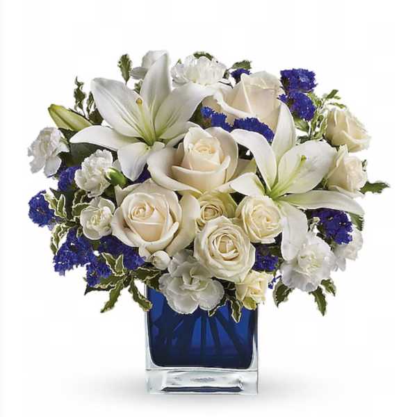 Compact white lilies, roses, and carnations with blue accents in a blue glass cube vase