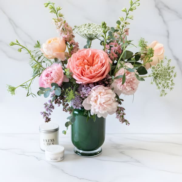 Pastel pink and peach flower arrangement in a dark green glass vase with two small white candle tins.