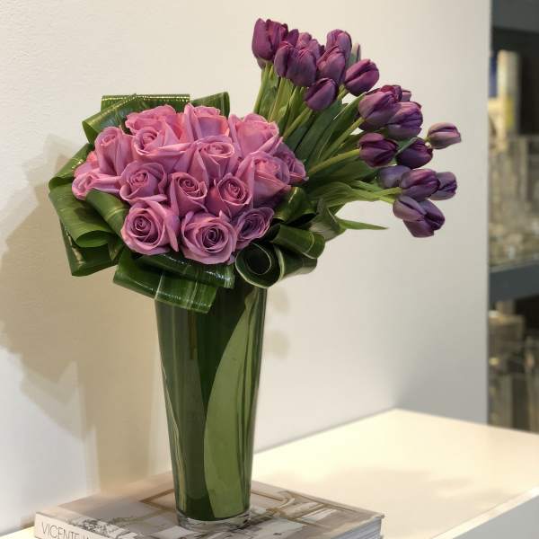 Tall vase arrangement of pink roses and purple tulips with folded green foliage.