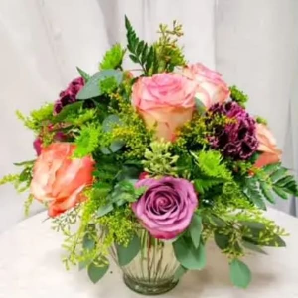 Round arrangement of peach and lavender roses with dark plum flowers in a clear glass vase