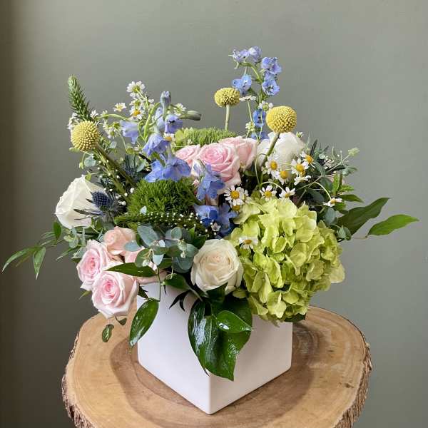 Mixed bouquet in a white square vase with pink roses, green hydrangea, and blue flowers