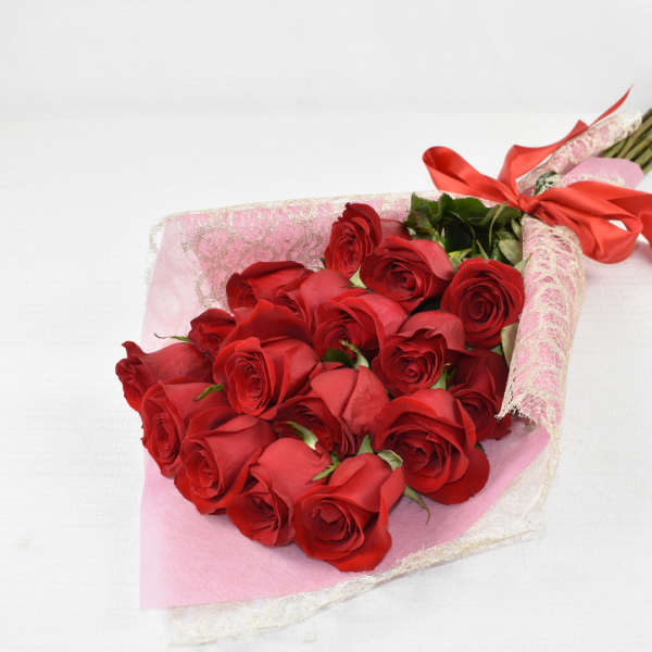 Hand-tied bouquet of long-stem red roses wrapped in pink lace paper with a red ribbon bow