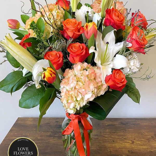 Tall mixed arrangement of orange roses, tulips, white lilies, and peach hydrangeas in a clear vase with orange ribbon