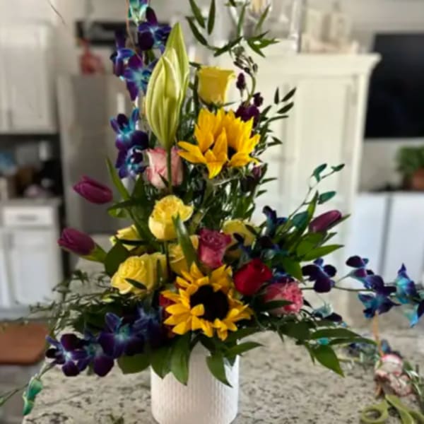 Tall bouquet with sunflowers, roses, lilies, and purple orchids in a white vase.