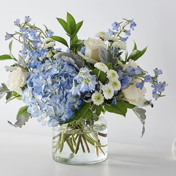 Low glass vase of blue hydrangea, white roses, white mums, and blue delphinium.