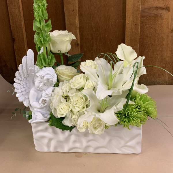 White floral arrangement with roses, lilies, and a cherub figurine in a white container