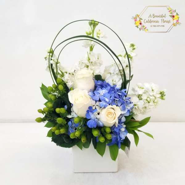 White and blue floral arrangement in a white box with arched handles