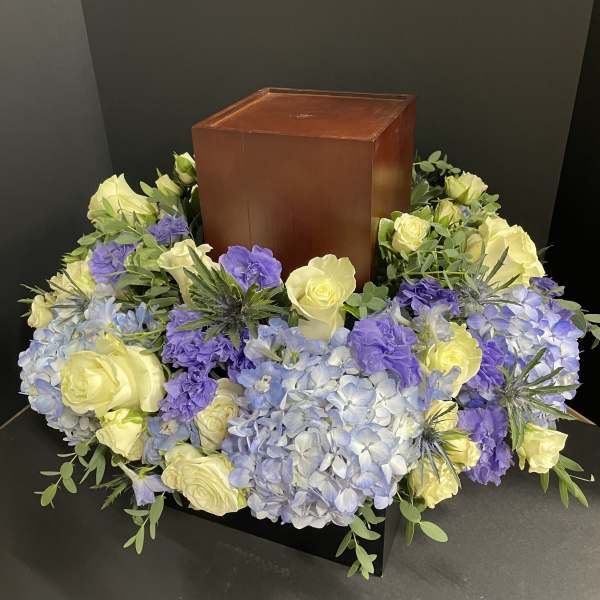 Floral wreath around a wooden urn with pale yellow roses and blue-purple hydrangeas