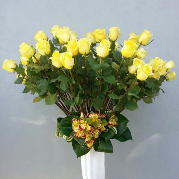 Tall arrangement of yellow roses in a white vase with a small orchid cluster at the base