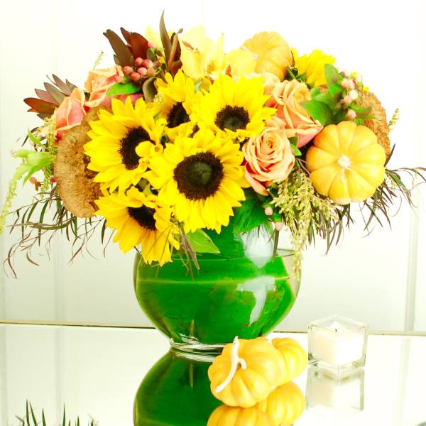 Sunflower and peach rose arrangement in a green glass vase with small pumpkins