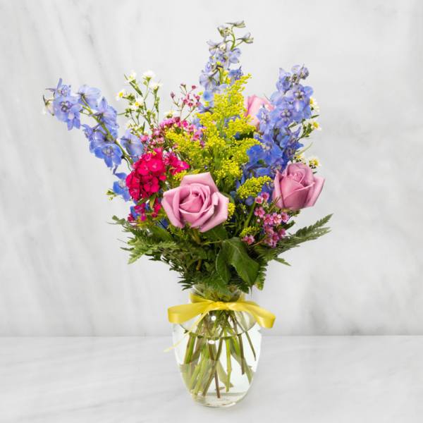 Mixed bouquet of pink roses, blue delphinium, and yellow filler in a glass vase