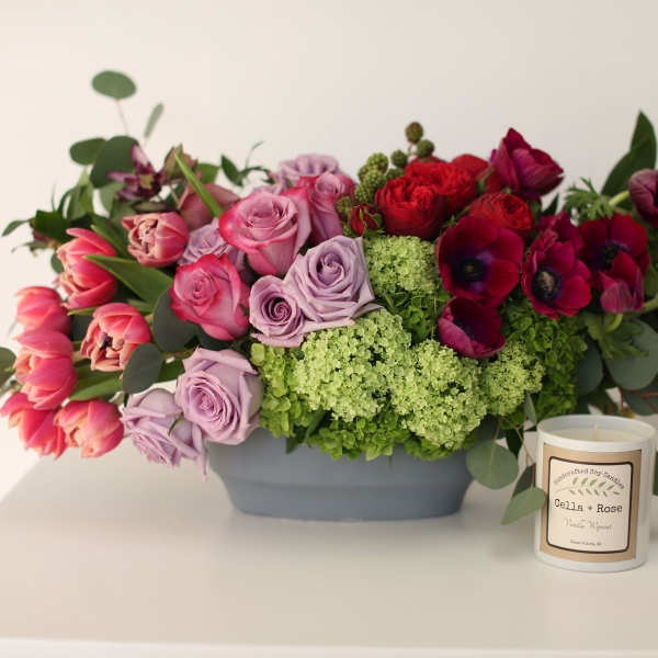 Mixed bouquet of roses, tulips, and anemones in a gray container beside a candle