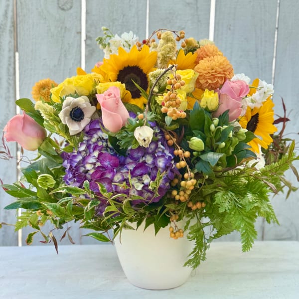Mixed bouquet of sunflowers, roses, hydrangea, and white blooms in a white vase