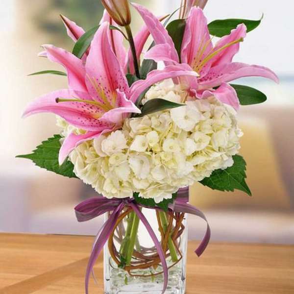 Pink lilies and white hydrangeas in a clear glass vase with a purple ribbon