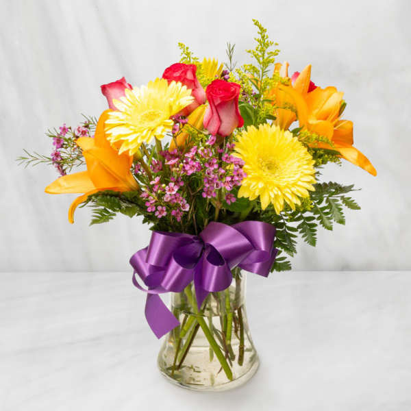 Bouquet of yellow daisies, orange lilies, and red roses in a glass vase with a purple ribbon