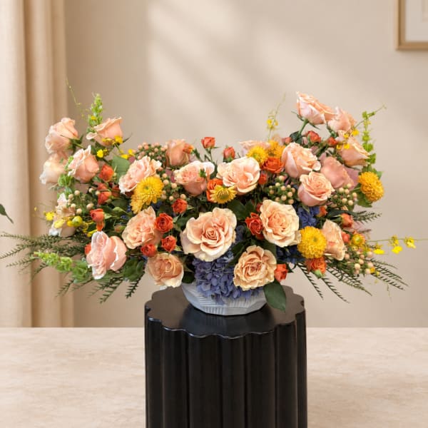 Peach and yellow floral arrangement in a white vase on a black pedestal
