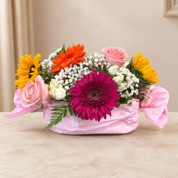 Mixed bouquet in a pink-wrapped basket with roses, gerberas, and sunflowers