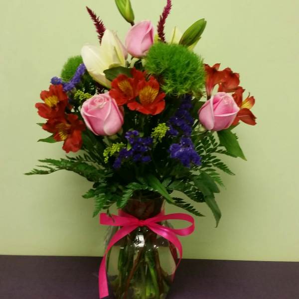 Mixed bouquet of pink roses, lilies, and red flowers in a glass vase with a pink ribbon.