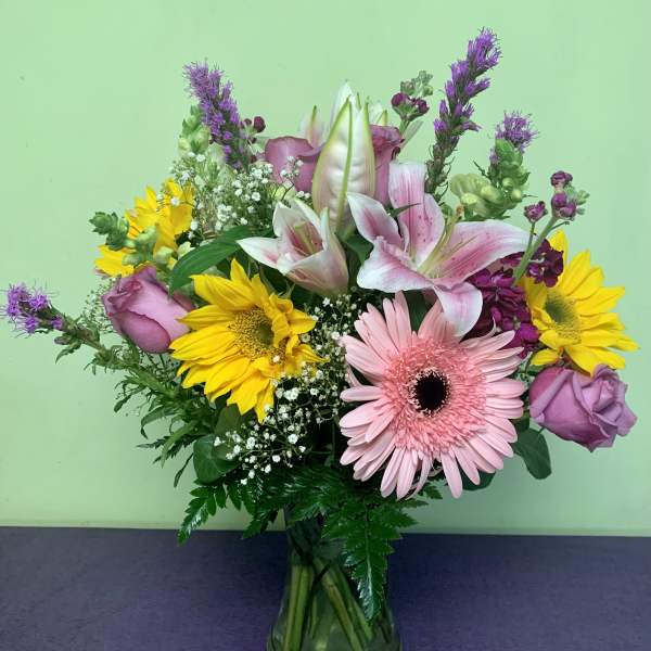 Mixed bouquet of pink lilies, roses, gerbera daisies, and sunflowers in a glass vase