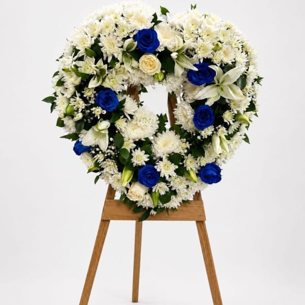Heart-shaped floral wreath of white flowers with blue roses on a wooden stand
