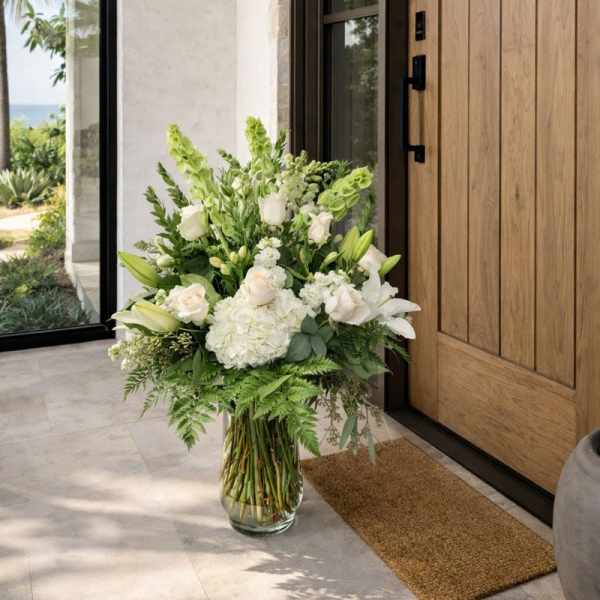 Tall white floral arrangement in a glass vase by a wooden door