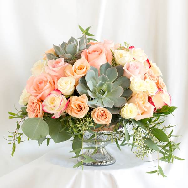 Peach and cream roses arranged with succulents in a glass pedestal vase