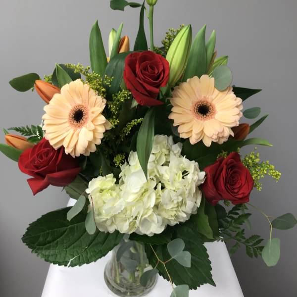Bouquet of red roses, peach gerbera daisies, white hydrangea, and lilies in a glass vase