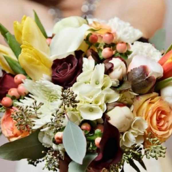 Hand-tied bouquet of ivory, peach, yellow, and burgundy flowers with berries, held by a person in white.