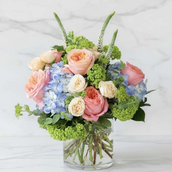 Low arrangement of peach and pink roses, cream roses, and blue hydrangeas in a clear glass vase.