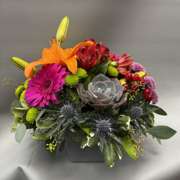 Colorful low arrangement with orange lilies, pink gerbera, succulents and mixed blooms in a gray square vase